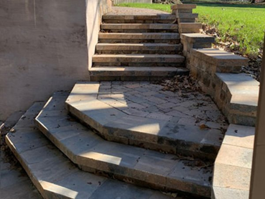 Stone paver patio with tiered retaining wall and multi-level steps leading up beside a house, with scattered leaves and a grassy yard in the background.