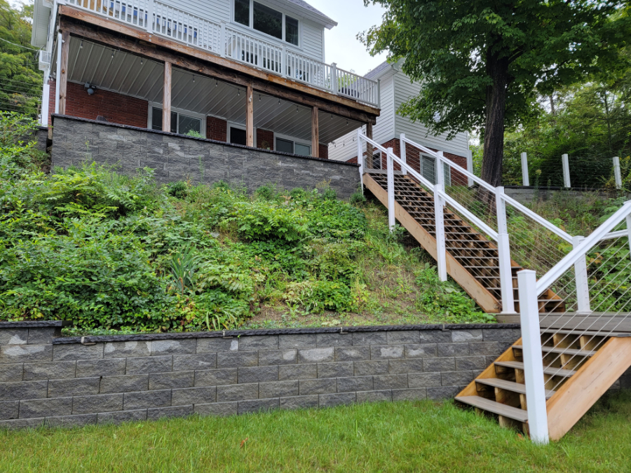 Backyard deck with wooden stairs, cable railing, and retaining wall on a sloped yard behind a residential home in Seneca Falls, NY