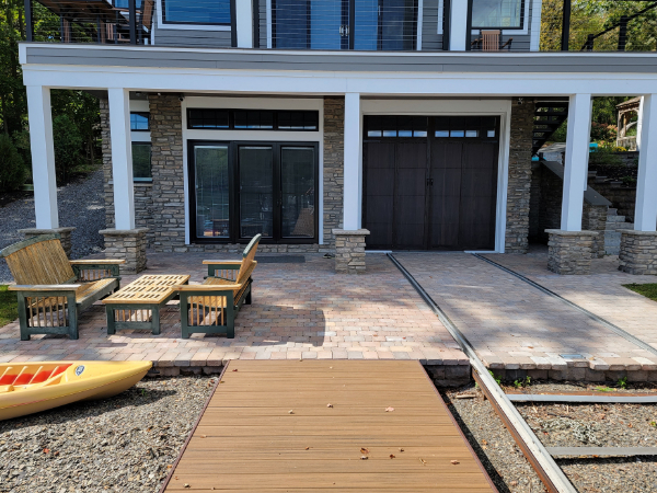 Waterfront paver patio with outdoor seating area in front of a covered structure, connected to a dock leading to the shoreline.