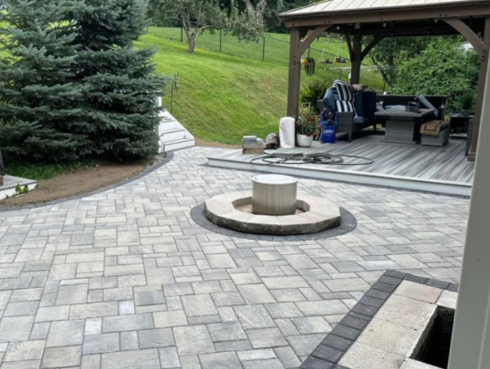 Backyard paver patio with a circular fire pit in the center and a covered seating area with outdoor furniture in the background in Geneva, NY.