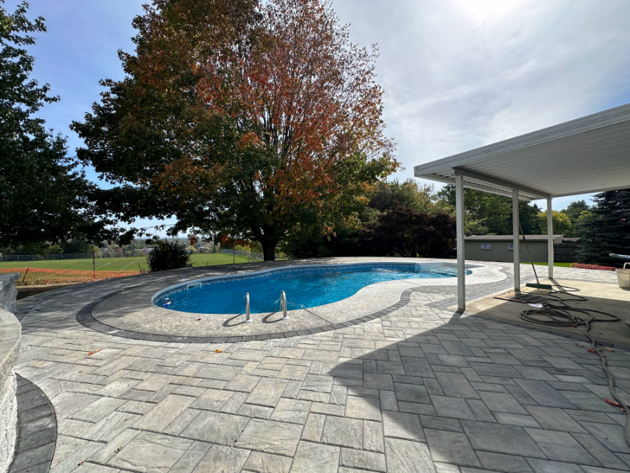 Curved in-ground swimming pool surrounded by gray paver patio with decorative border, next to a covered patio and landscaped yard.