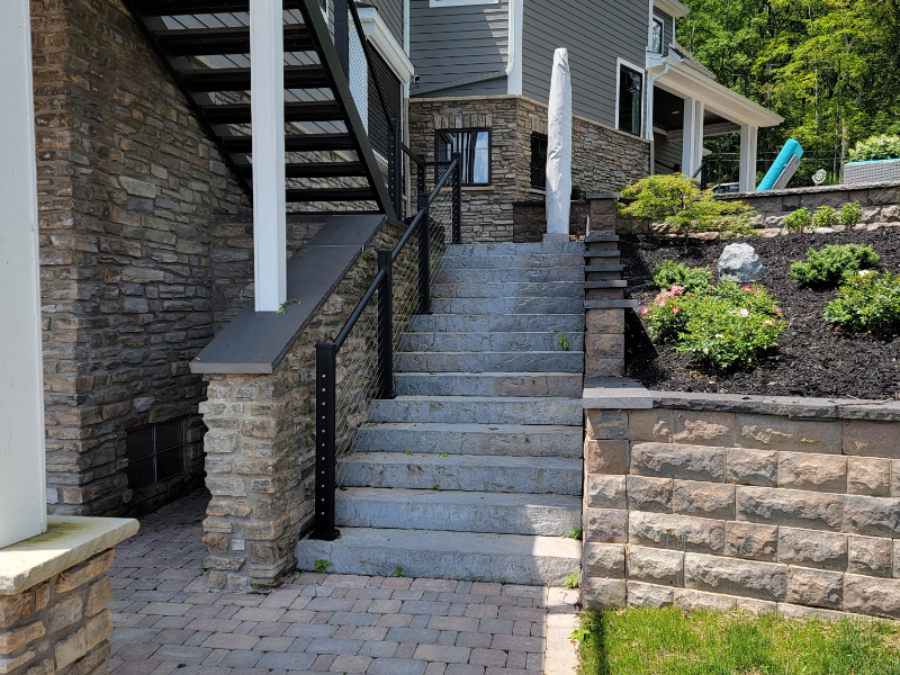 Stone staircase with black metal railing leading up alongside a home, bordered by retaining walls and landscaped garden beds.