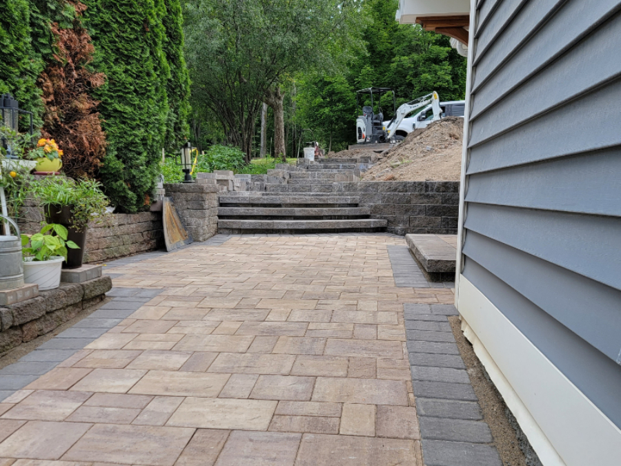 Newly installed Concrete paver patio with multiple retaining walls and wide steps leading up a sloped backyard beside a house in Seneca Falls, NY.