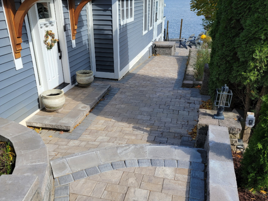 Brick paver walkway and small patio along the side of a blue house, with curved stone steps and edging, planters by the door, a retaining wall with lantern-style lighting, and a view leading down toward the waterfront.