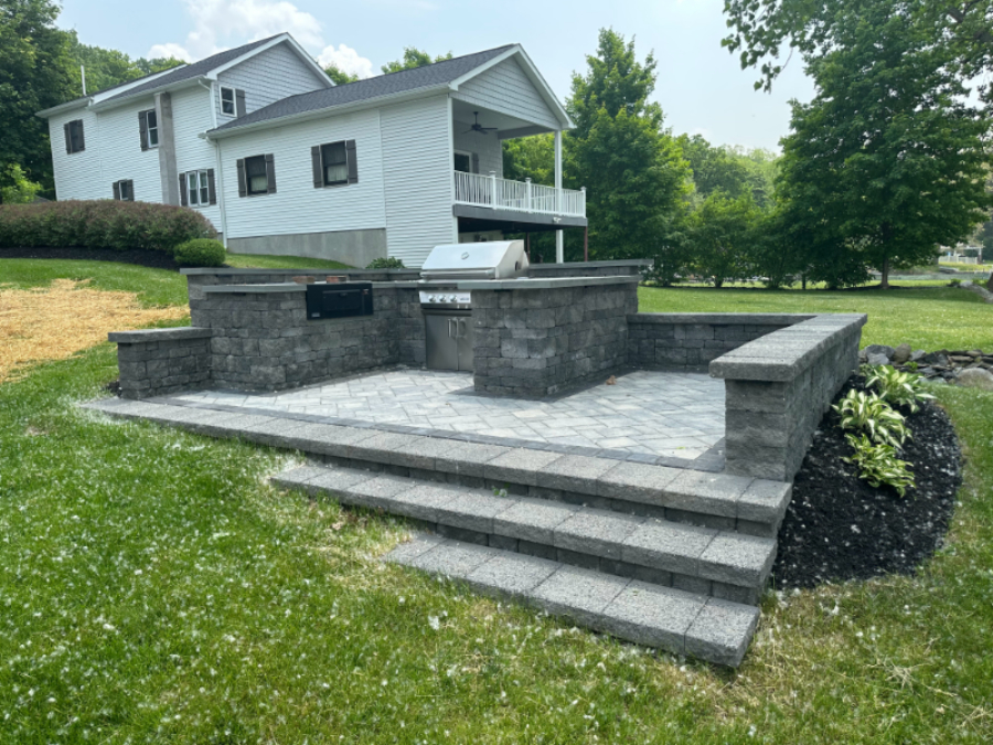 Raised paver patio with a built-in outdoor kitchen and grill station, surrounded by a block retaining wall in an open backyard in Penn Yan, NY.