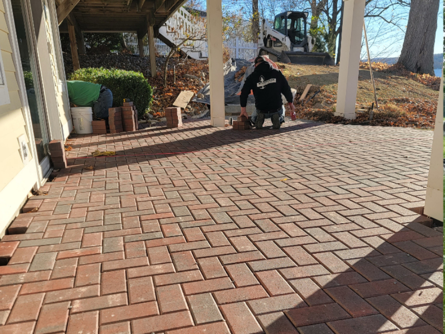 Contractor installing a brick paver patio under a covered structure beside a home, carefully placing pavers with tools and materials arranged nearby in Geneva, NY.