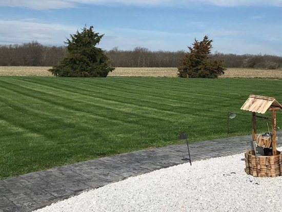 Wide, freshly mowed lawn with striped grass patterns bordered by a curved stone edge and gravel area in a rural setting in Geneva, NY.