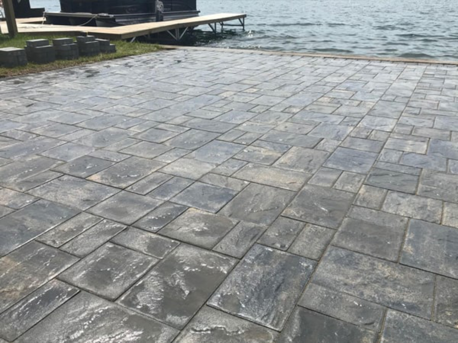 Freshly installed gray paver patio with smooth, uniform surface set along a waterfront, with a wooden dock and calm lake visible in the background.